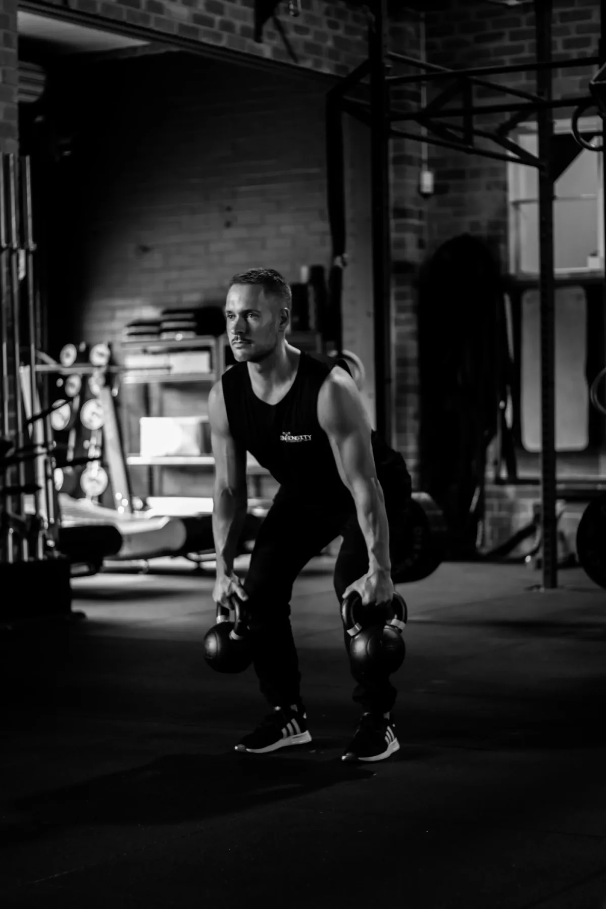 Black and white image of Alex Bastianon in half-squat position, inside a gym, holding a kettlebell weight in each hand